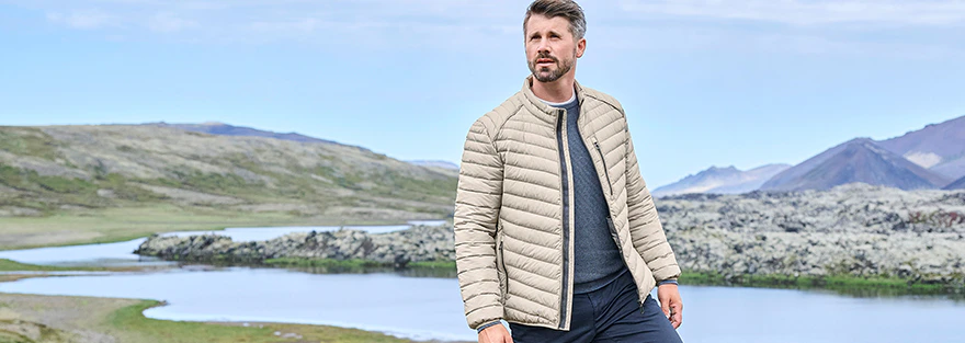 Thore Schölermann steht in hügeliger Landschaft mit See im Hintergrund und trägt eine beige gesteppte Jacke, dunkle Hose und helle Schuhe