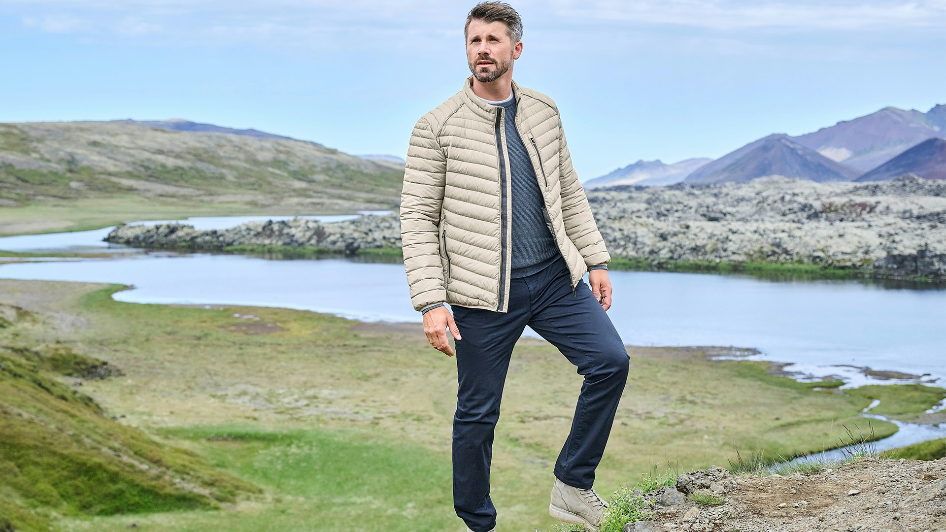 Thore Schölermann steht in hügeliger Landschaft mit See im Hintergrund und trägt eine beige gesteppte Jacke, dunkle Hose und helle Schuhe