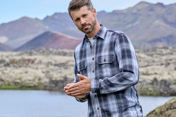 Thore Schölermann steht in hügeliger Landschaft mit See im Hintergrund und trägt ein kariertes Langarmhemd und Jeans