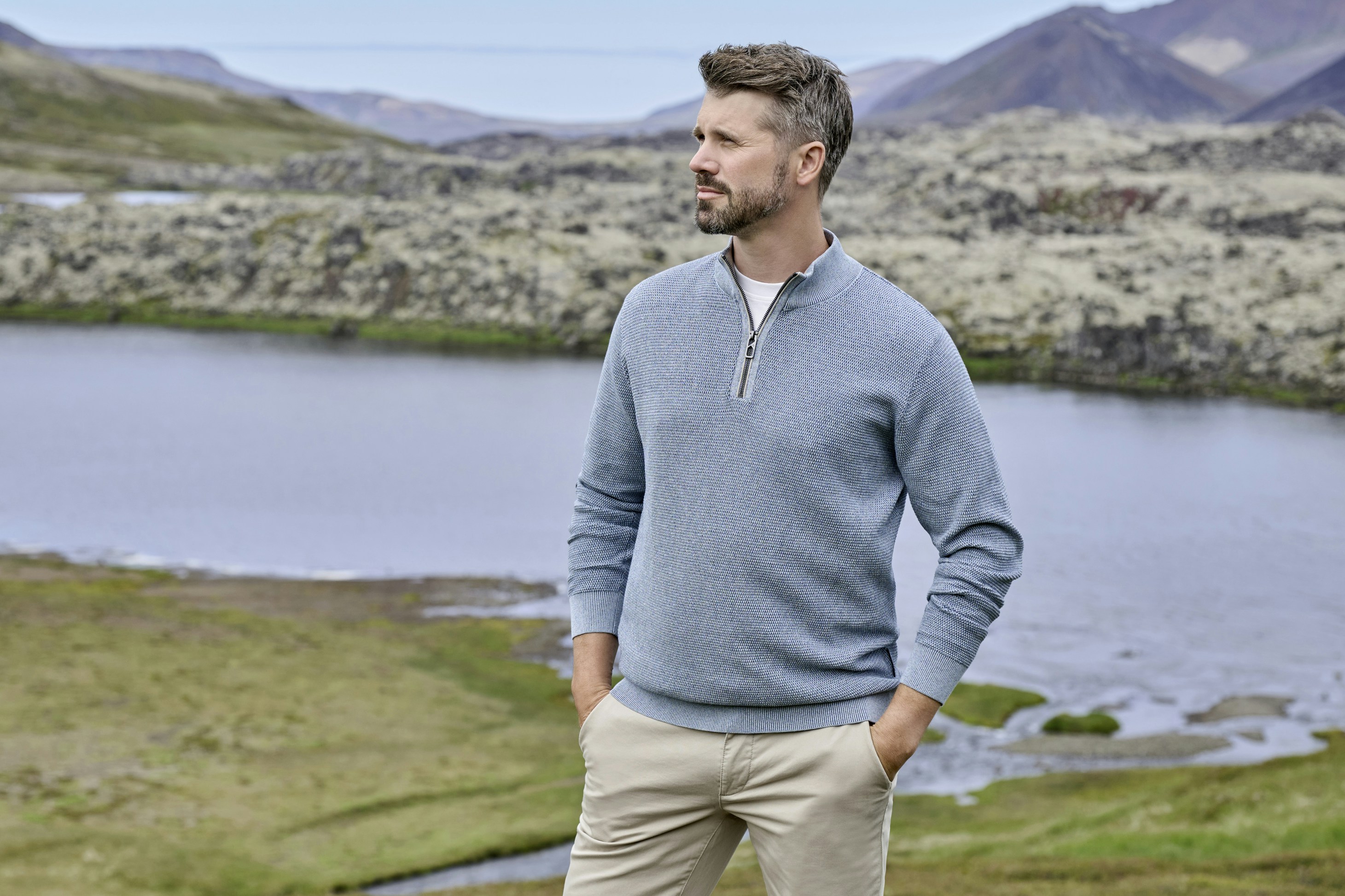 Thore Schölermann steht vor See und felsiger Landschaft und trägt einen hellblauen Pullover mit Reißverschluss am Kragen und helle Hose