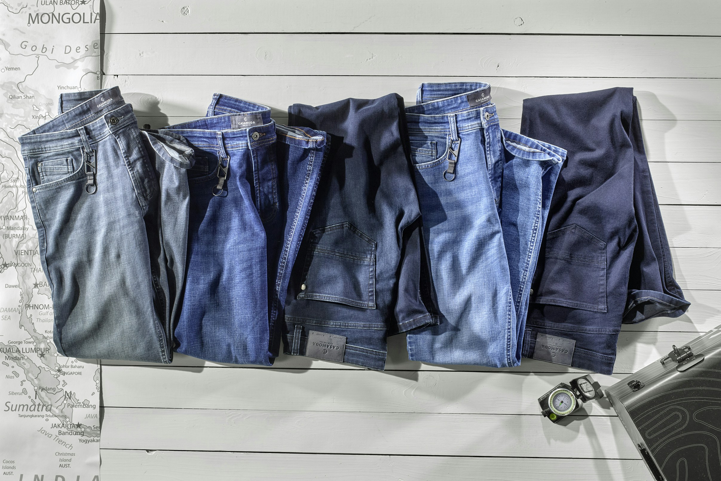 Fünf gefaltete Jeans in verschiedenen Blau- und Dunkelblautönen liegen parallel auf weißem Holzboden. Jede Jeans ist mit einem Metall-Clip an der Gürtelschlaufe versehen. Kartenausschnitt links, Koffer und Kompass rechts