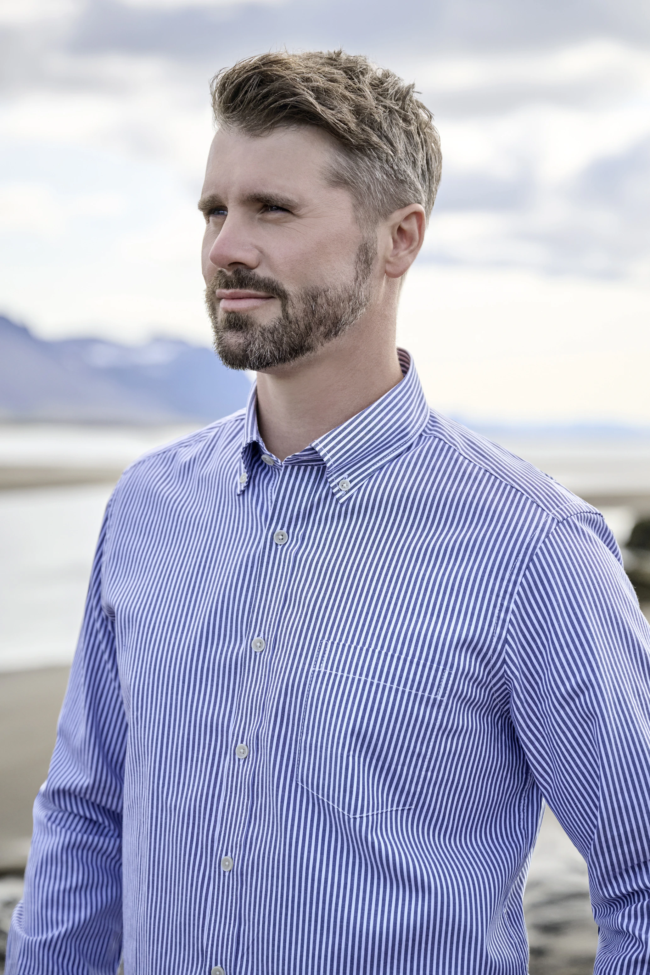 Thore Schölermann steht im Freien vor Wasser- und Berglandschaft und trägt ein blau-weiß gestreiftes Hemd