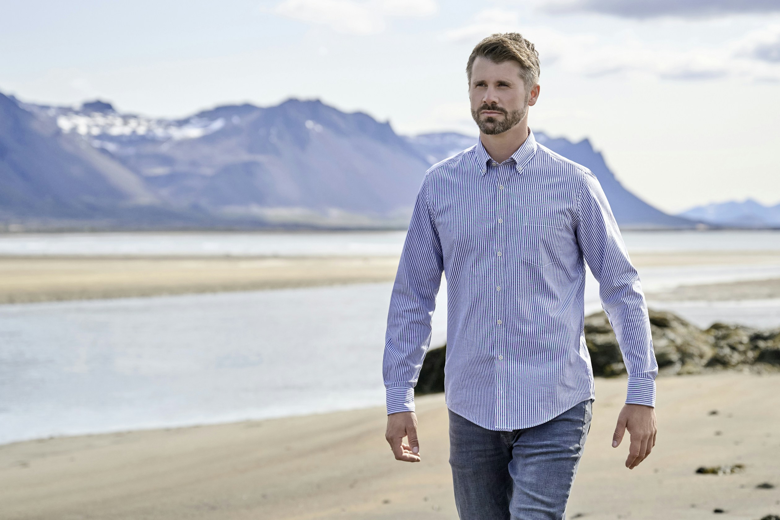 Thore Schölermann läuft am Strand mit Wasser und Bergen im Hintergrund und trägt ein blau-weiß gestreiftes Langarmhemd und Jeans