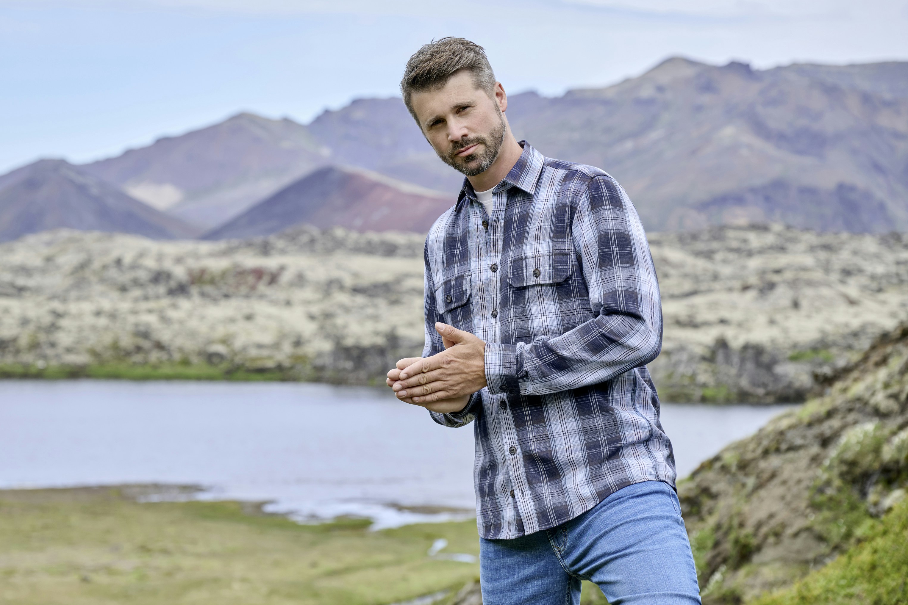 Thore Schölermann steht in hügeliger Landschaft mit See im Hintergrund und trägt ein kariertes Langarmhemd und Jeans