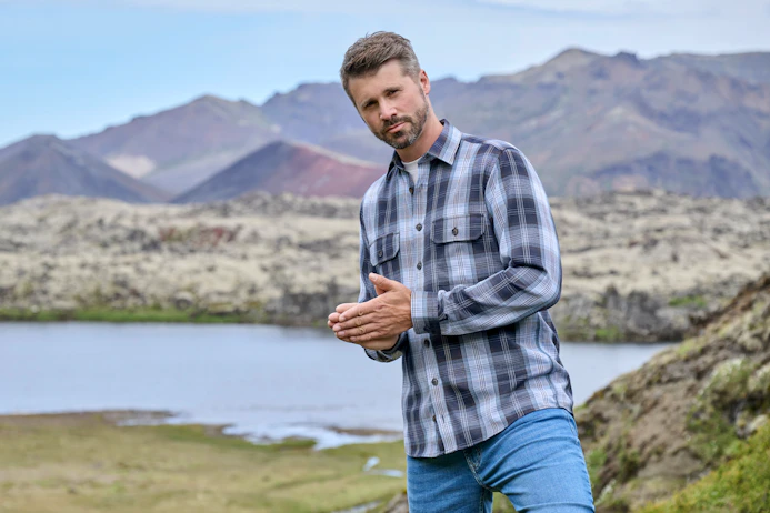 Thore Schölermann steht in hügeliger Landschaft mit See im Hintergrund und trägt ein kariertes Langarmhemd und Jeans
