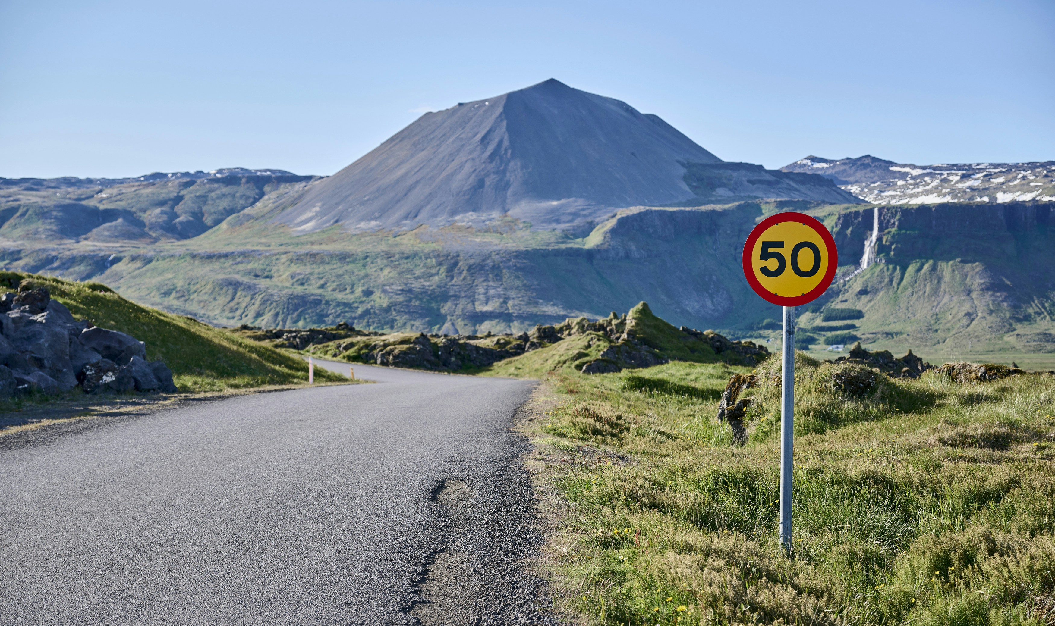 Leere Straße führt durch hügelige Landschaft mit Bergen, rechts steht ein rundes Verkehrsschild mit der Zahl 50