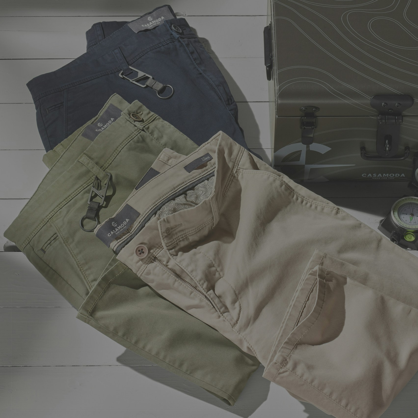 Drei gefaltete Hosen in Dunkelblau, Olivgrün und Beige liegen übereinander. Sichtbar sind Bund, Knopf, Reißverschluss und Gesäßtaschen. Rechts steht ein geschlossener Koffer, daneben ein Kompass.