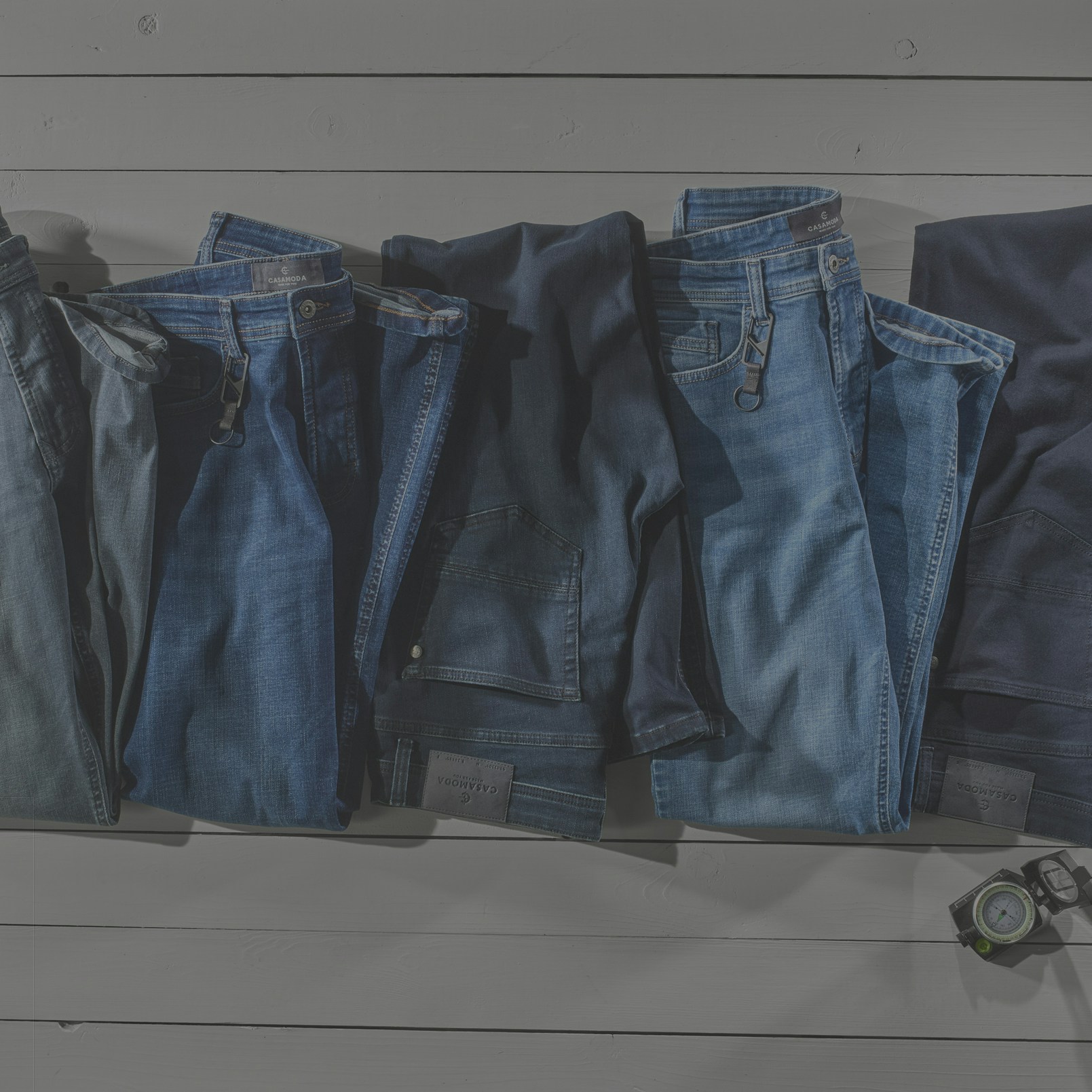 Fünf gefaltete Jeans in verschiedenen Blau- und Dunkelblautönen liegen parallel auf weißem Holzboden. Jede Jeans ist mit einem Metall-Clip an der Gürtelschlaufe versehen. Kartenausschnitt links, Koffer und Kompass rechts