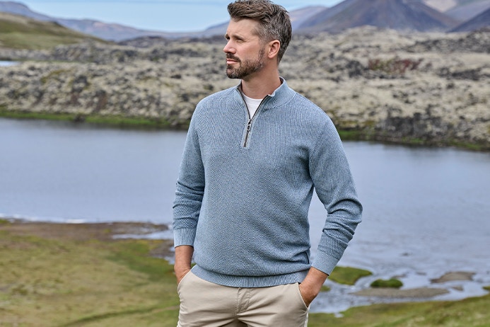 Thore Schölermann steht vor See und felsiger Landschaft und trägt einen hellblauen Pullover mit Reißverschluss am Kragen und helle Hose
