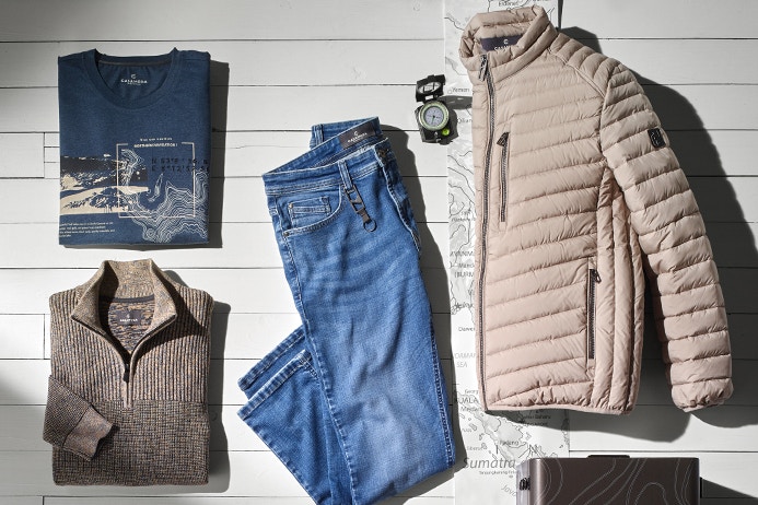 Flach liegende Produktkombination: links ein gefaltetes blaues T-Shirt mit Print, in der Mitte liegt eine gefaltete blaue Jeans. Rechts eine beige gesteppte Jacke mit Reißverschluss und Seitentaschen. Hintergrund ist weißer Holzboden mit Kartenelementen, Kompass und Koffer.