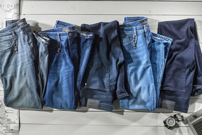 Fünf gefaltete Jeans in verschiedenen Blau- und Dunkelblautönen liegen parallel auf weißem Holzboden. Jede Jeans ist mit einem Metall-Clip an der Gürtelschlaufe versehen. Kartenausschnitt links, Koffer und Kompass rechts