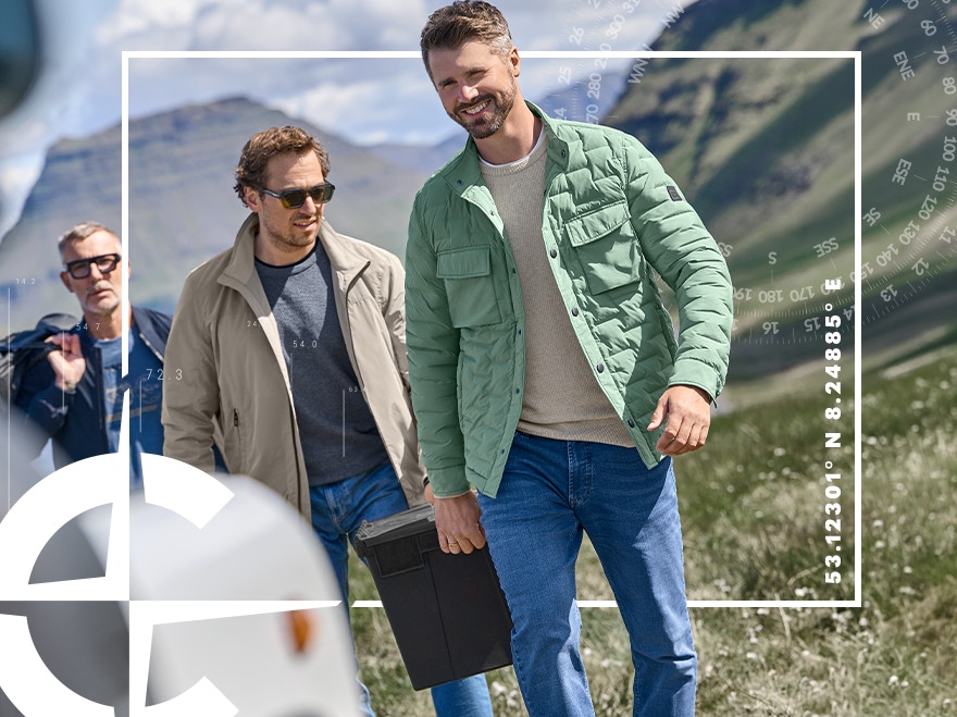 Thore Schölermann steht im Vordergrund in grüner Steppjacke, hellem Pullover und Jeans, neben ihm geht ein weiterer Mann in heller Jacke und Jeans durch eine bergige Landschaft