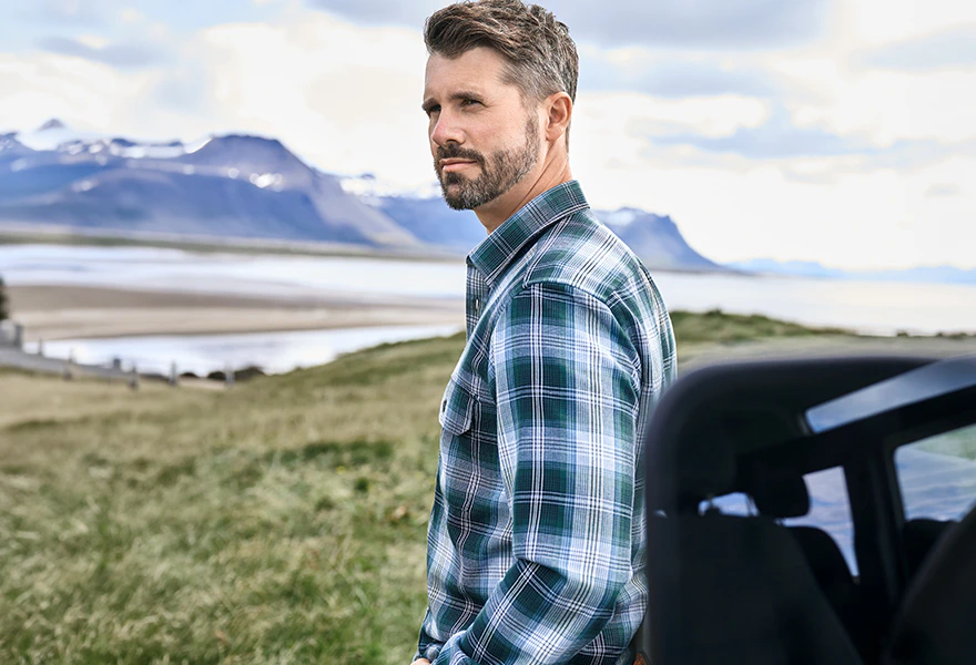 Thore Schölermann steht im Profil vor einer Landschaft mit Wasser und Bergen und trägt ein kariertes Hemd in Blau- und Grüntönen