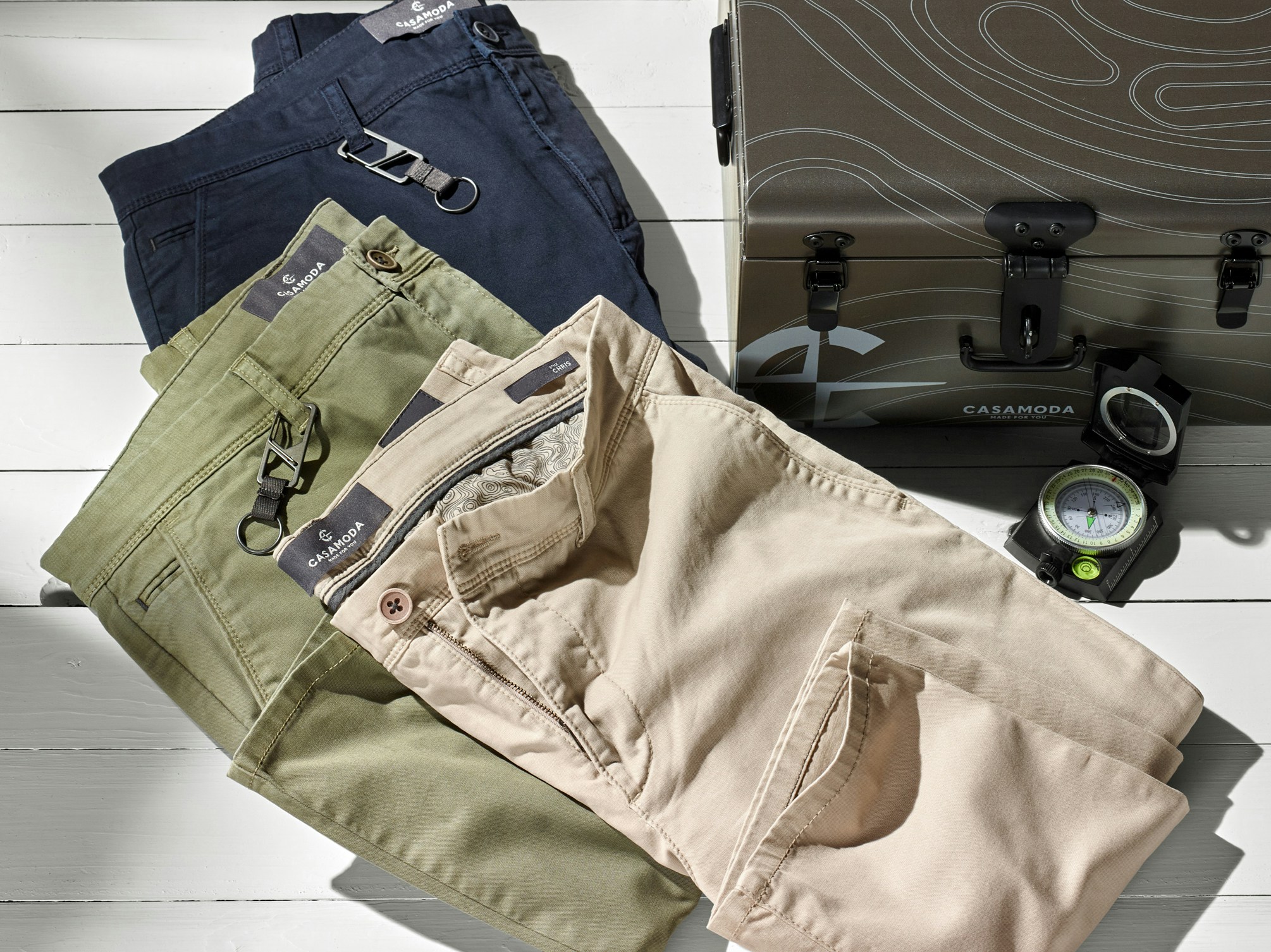 Mehrere gefaltete Hosen in Beige, Grün und Dunkelblau liegen auf weißem Holzboden. Rechts stehen ein Koffer und ein Kompass.