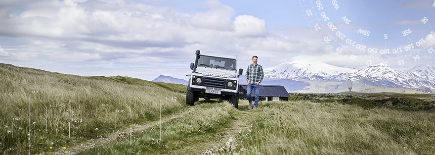 Thore Schölermann geht über eine Wiese neben einem weißen Geländewagen, im Hintergrund sind Berge und ein Gebäude zu sehen