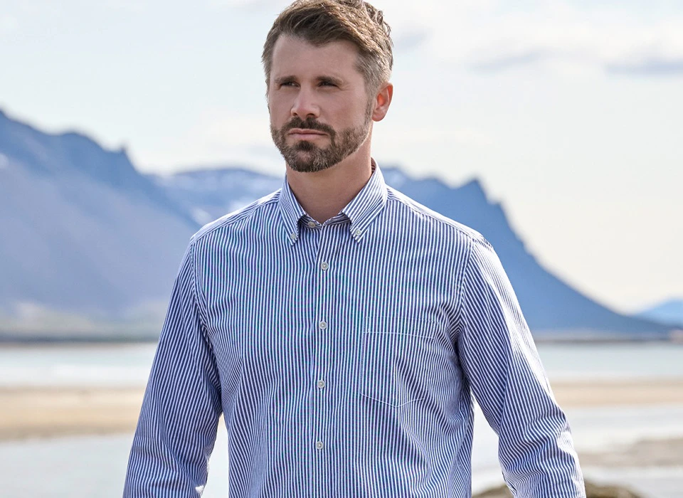 Thore Schölermann läuft am Strand mit Wasser und Bergen im Hintergrund und trägt ein blau-weiß gestreiftes Langarmhemd und Jeans