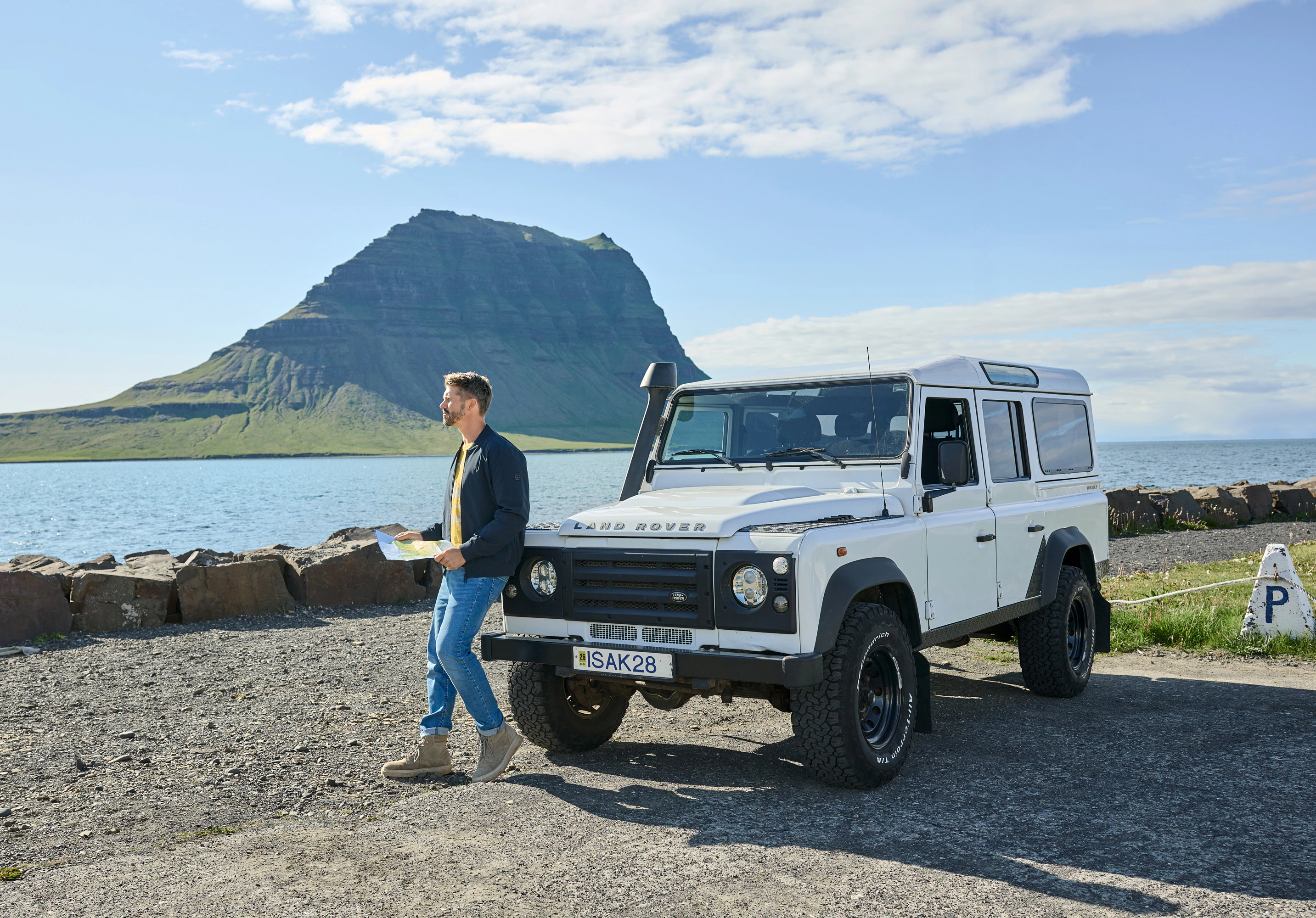 CASAMODA Herrenmode: Mann mit Jacke und Jeans neben Land Rover am Meer, Bergkulisse im Hintergrund