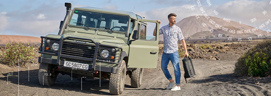 Herren Kurzarmhemd mit floralem Muster, Jeans und weißen Sneakern vor Geländewagen in vulkanischer Landschaft