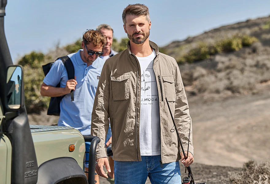 Herren Leichtjacke in Beige über Print-T-Shirt und Jeans bei einem Outdoor-Ausflug mit Geländewagen