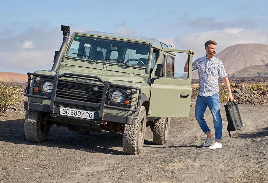 Herren Kurzarmhemd mit floralem Muster, Jeans und weißen Sneakern vor Geländewagen in vulkanischer Landschaft