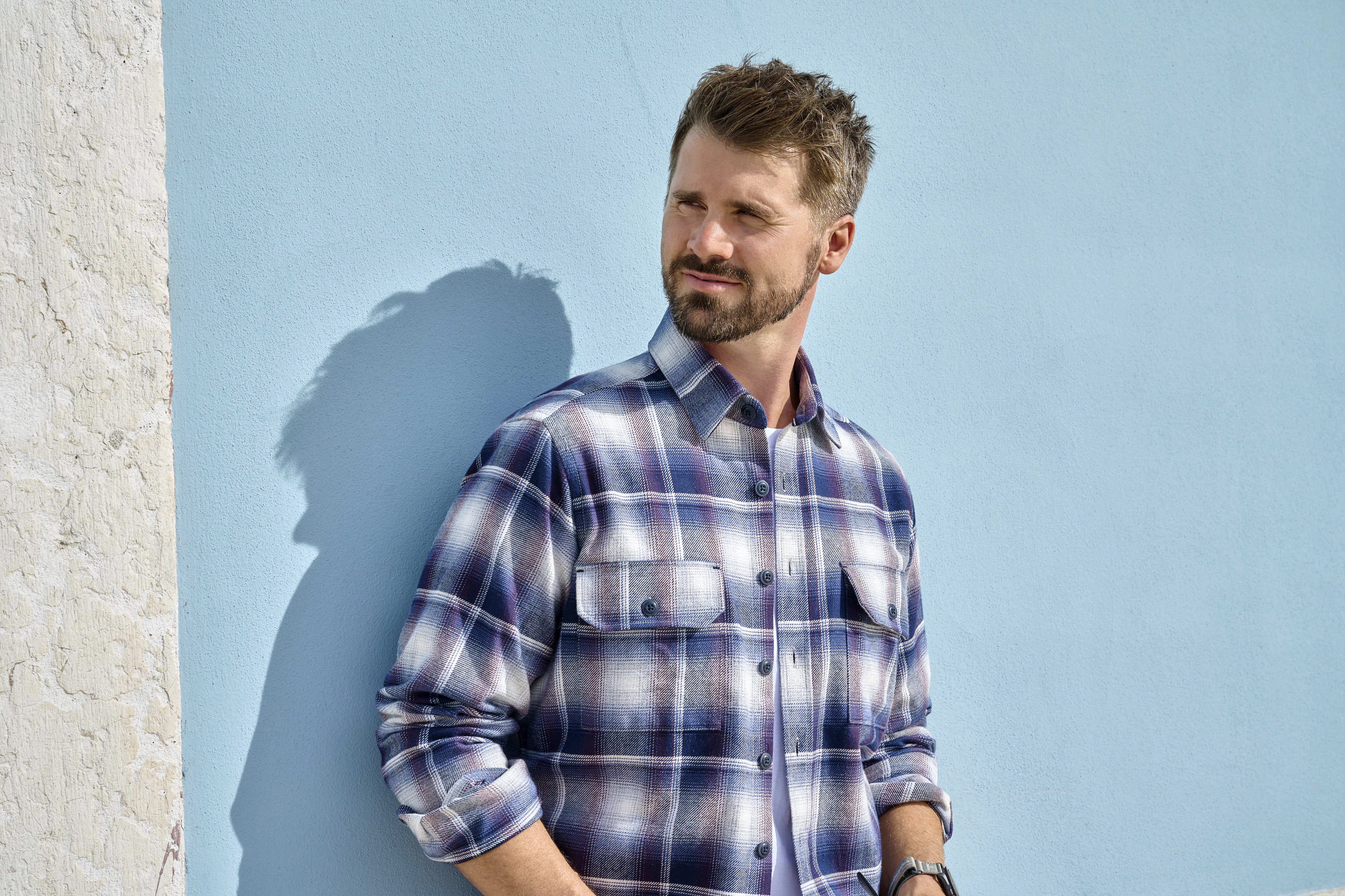 Thore Schölermann posiert vor einer blauen Wand mit einem karierten Hemd von Casmoda