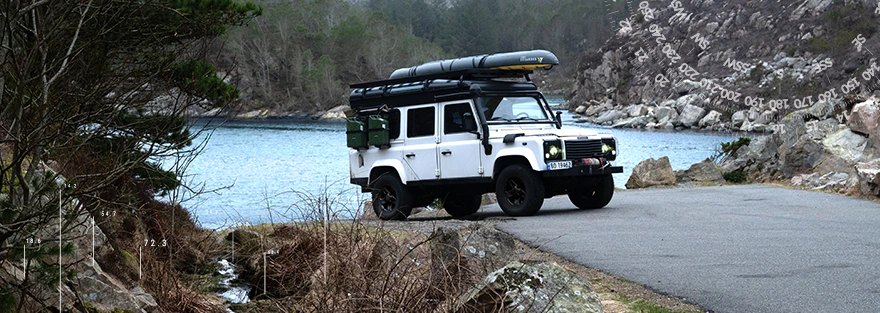 Weißer Geländewagen mit Dachgepäck steht an einer felsigen Küstenstraße direkt am Wasser.