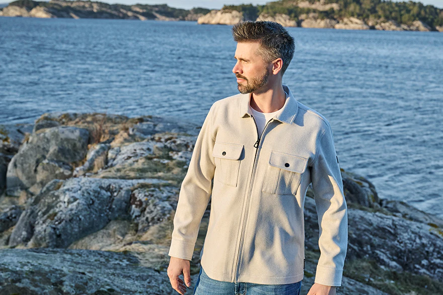 Thore Schölermann trägt eine beige Hemdjacke mit Brusttaschen über weißem T-Shirt an einer felsigen Küste.