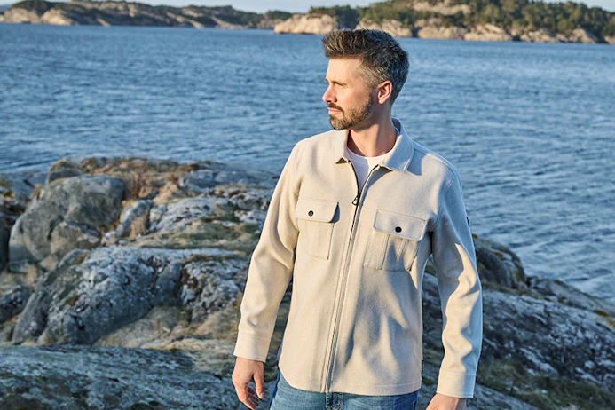 Thore Schölermann trägt eine beige Hemdjacke mit Brusttaschen über weißem T-Shirt an einer felsigen Küste.