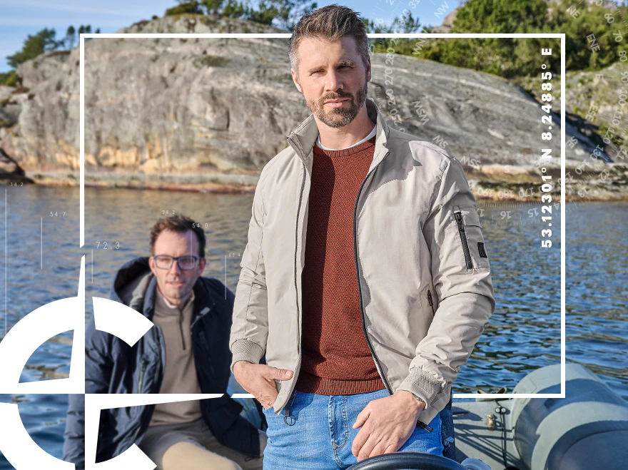 Thore Schölermann trägt eine beige Jacke über rostbraunem Pullover und blauer Jeans, begleitet von einem Mann im Boot.