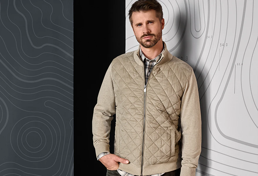 Thore trägt eine beige CASAMODA Herren-Steppjacke mit Rautensteppung und Strickärmeln, kombiniert mit einem karierten Hemd und dunkler Hose.