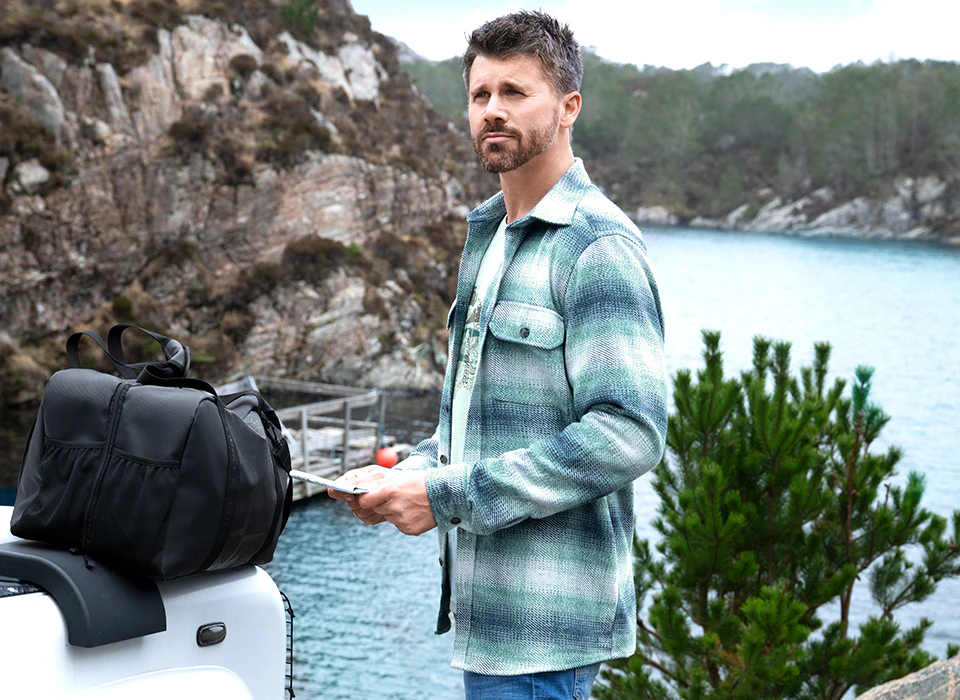 Thore Schölermann trägt ein grün-blau kariertes Overshirt über hellem T-Shirt vor einer felsigen Küstenlandschaft.