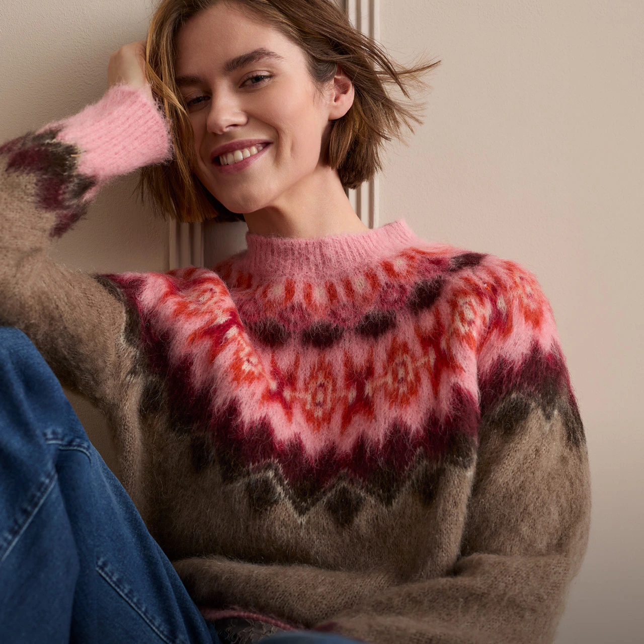 Model trägt einen braunen Norweger-Strickpullover mit pink-rotem Muster und blauer Jeans.