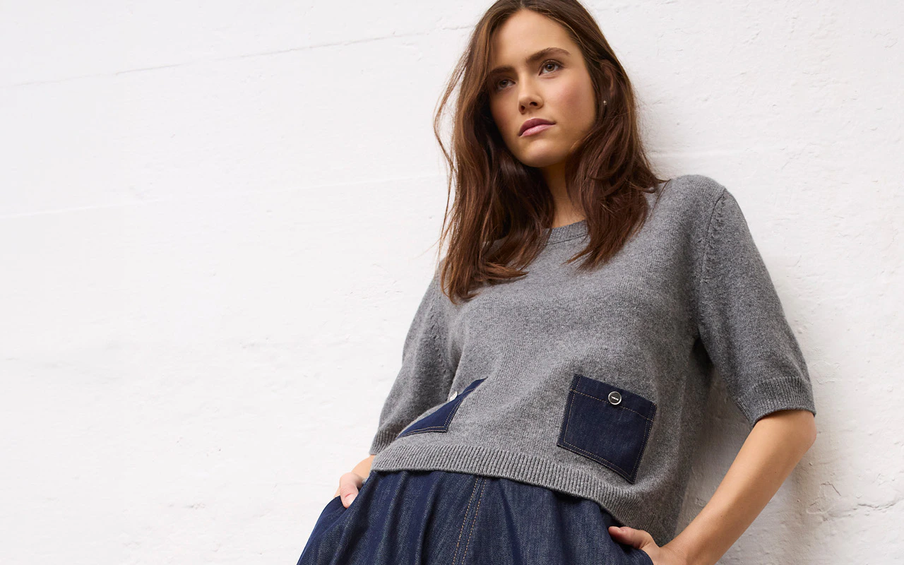 Model trägt einen grauen Strickpullover mit kurzen Ärmeln und aufgesetzten Denim-Taschen zu einer dunkelblauen Hose.