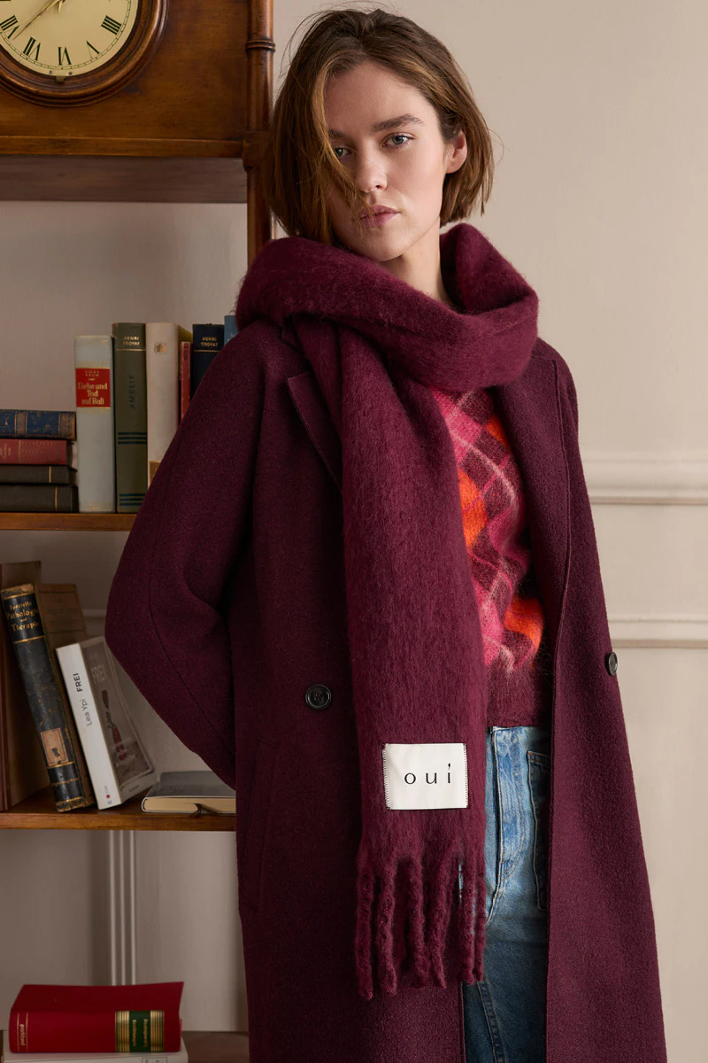 The model is wearing a burgundy coat with a matching scarf, a patterned jumper and jeans.