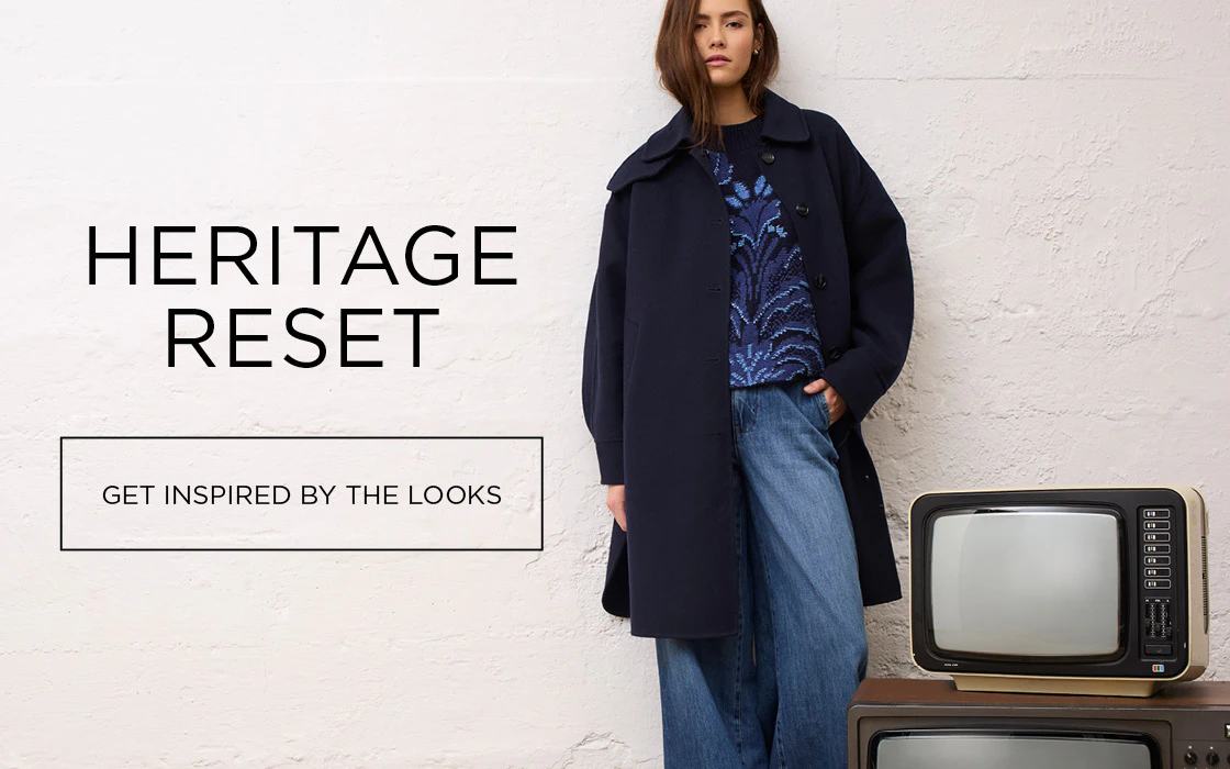 The model is wearing a dark blue coat, a patterned jumper and loose-fitting jeans in front of a white brick wall, next to two retro televisions.