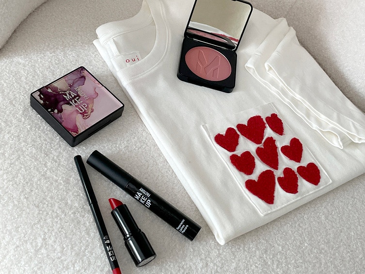 Ein weißes T-Shirt mit roten Herzmotiven liegt gefaltet auf einer Oberfläche. Darauf und daneben liegen Make-up-Produkte wie Rouge, Lippenstift, Eyeliner und eine Lidschattenpalette.