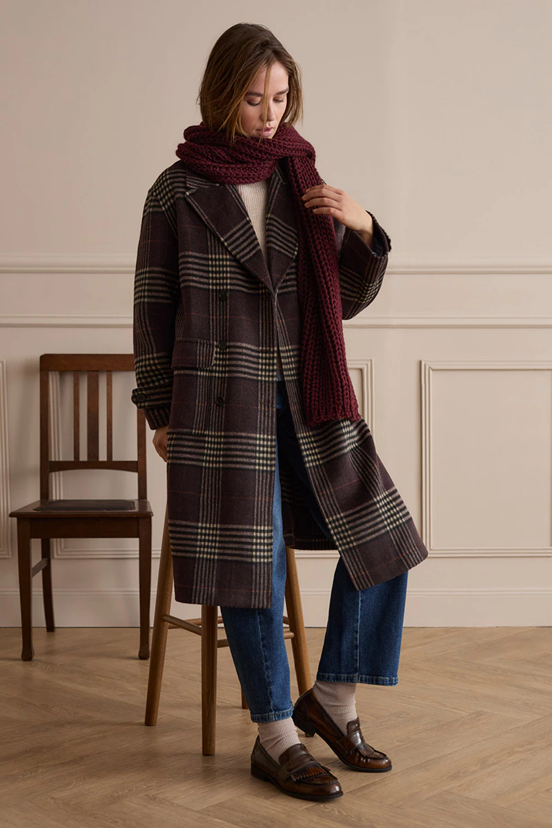 The model is wearing a dark brown checked coat with a burgundy scarf, jeans and brown loafers.