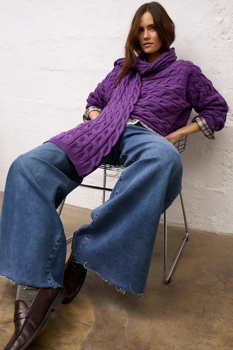A woman wearing a knitted purple jumper and a matching scarf, with jeans.