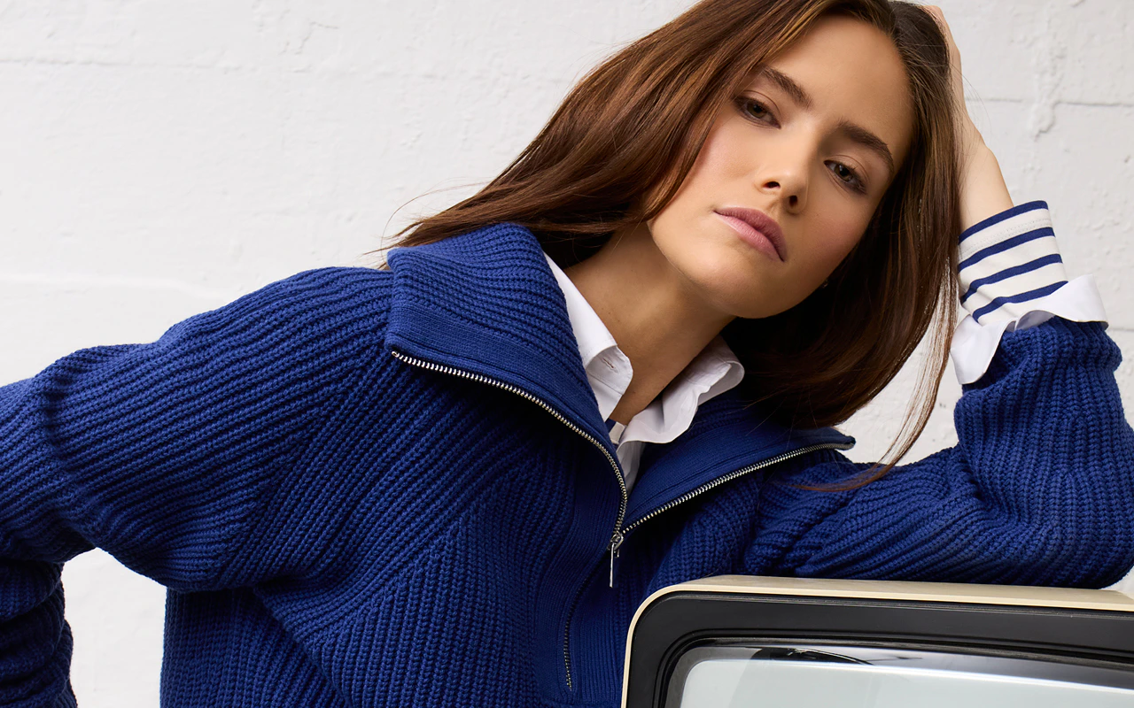 Alternativtext:
Frau im blauen Strickpullover mit Reißverschluss und weißem Hemd mit gestreiften Manschetten, lässig an einem Retro-Fernseher gelehnt.