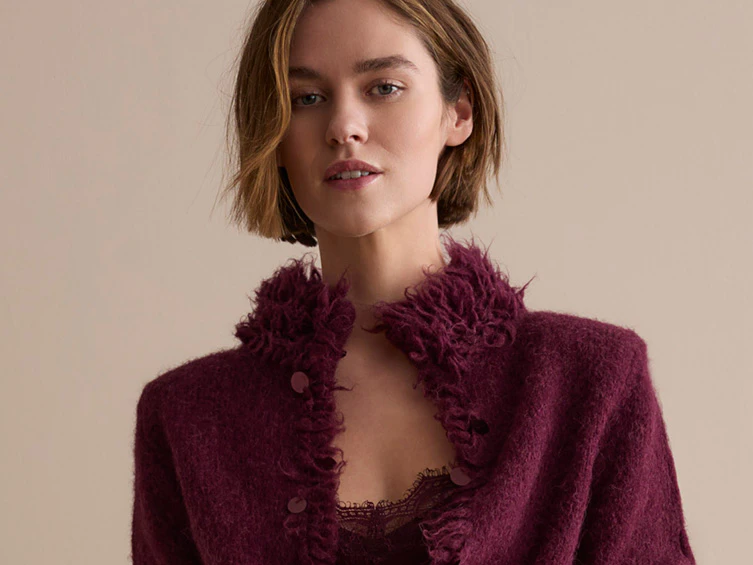 The model is wearing a burgundy cardigan with fringed detailing and a matching top, paired with a dark red leather midi skirt.