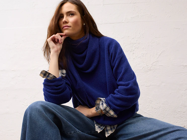 The model is wearing a blue jumper with a wide collar, a checked blouse underneath, and loose-fitting jeans from the Oui Heritage Reset collection.