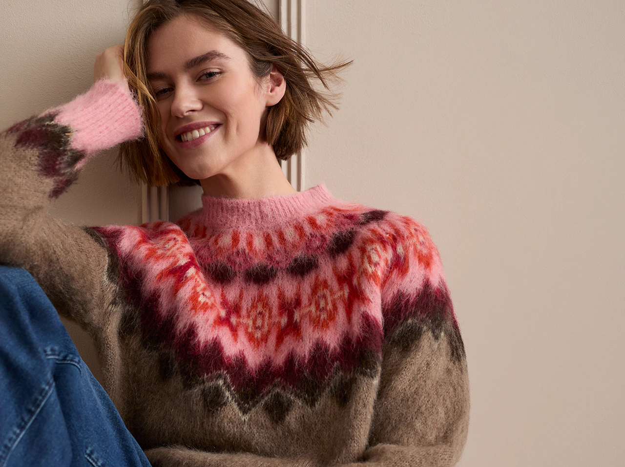 Model trägt einen braunen Norweger-Strickpullover mit pink-rotem Muster und blauer Jeans.