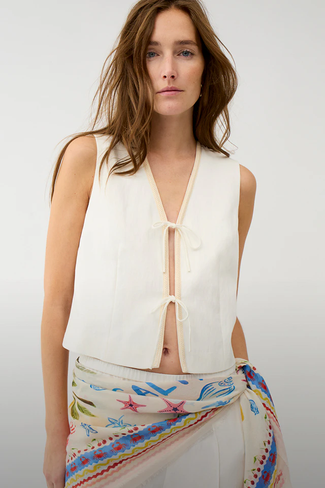 A woman is wearing a white vest top with a printed scarf tied around her waist and white trousers.