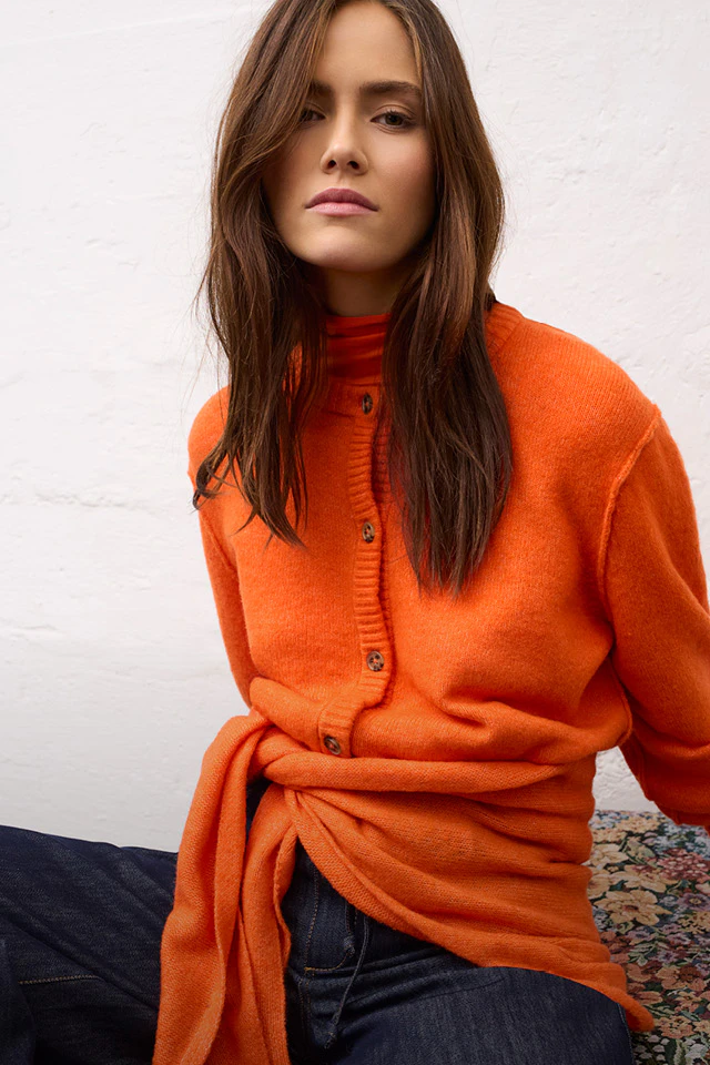 Model trägt eine orangene Strickjacke, ein oranges Dreieckstuch und Jeans.
