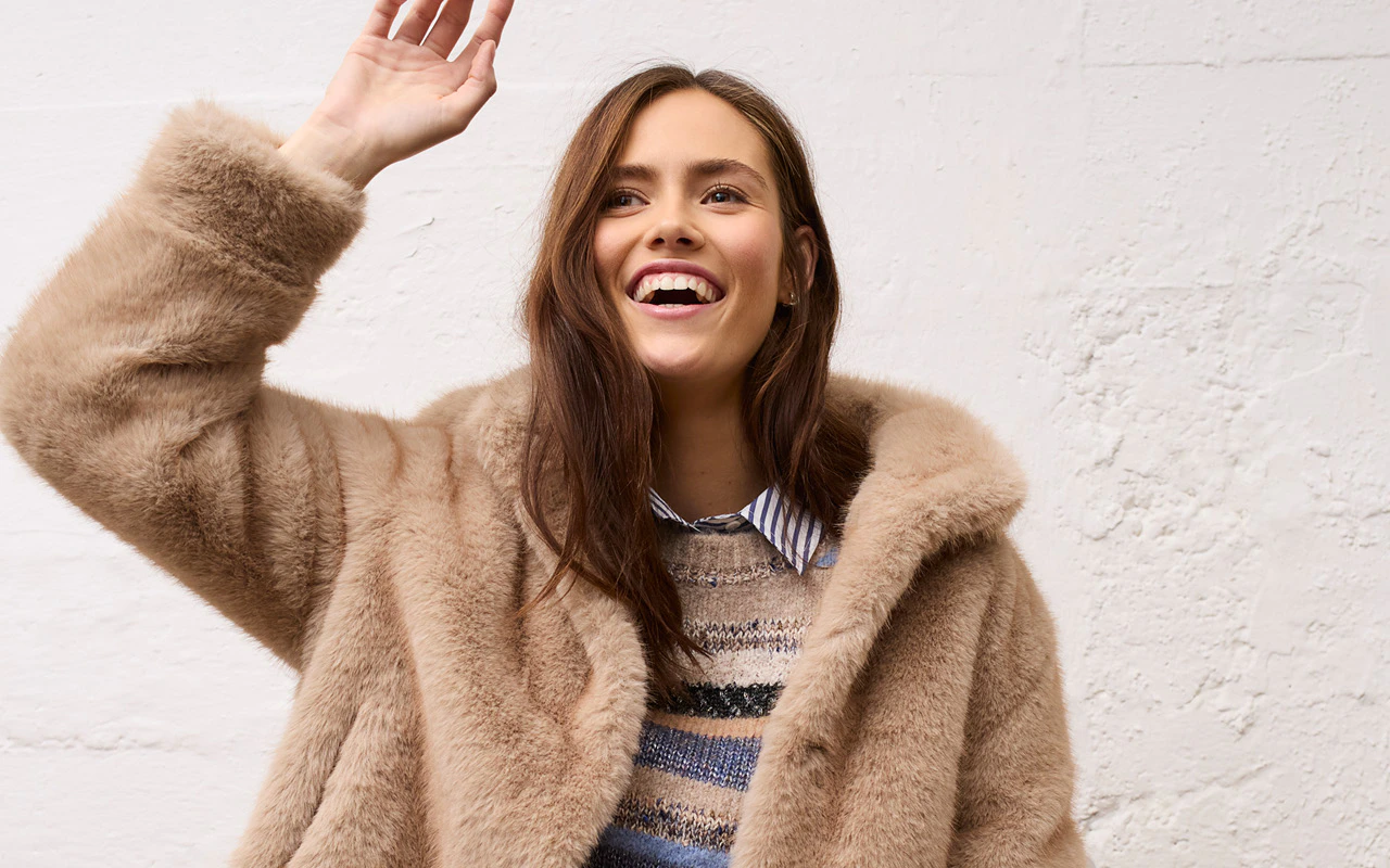 The model is wearing a beige faux fur coat over a patterned jumper with a striped shirt collar.