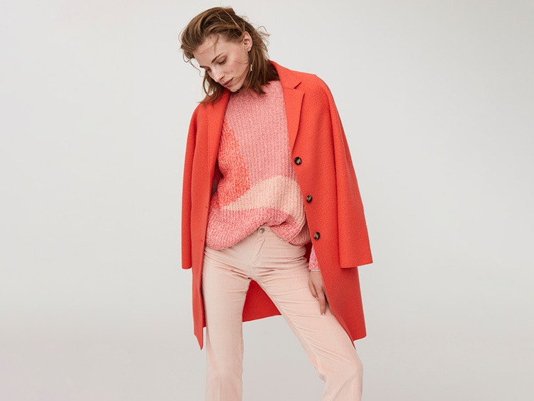 Eine Frau trägt einen leuchtend roten Mantel über einem rosafarbenen Pullover und einer hellrosa Hose.