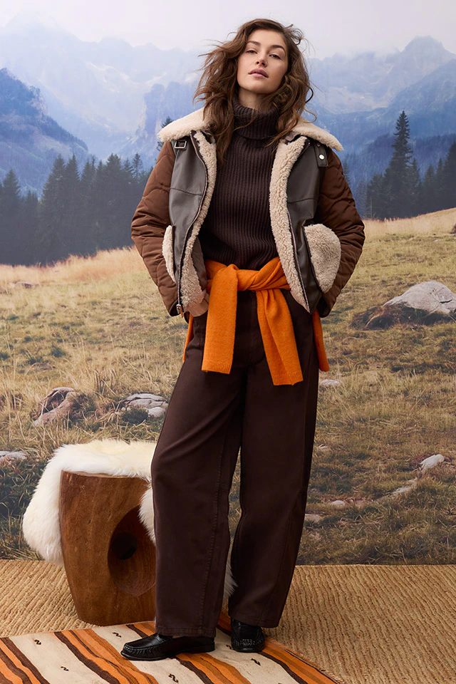 Damenmode im Herbst: Model trägt eine braune Jacke mit hellem Teddyfell-Besatz über einem dunkelbraunen Strickpullover. Ein orangefarbener Pullover ist um die Taille gebunden. 
