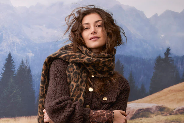 Model trägt eine dunkelbraune Strickjacke mit goldfarbenen Knöpfen, einen Leo-Schal und ein gemustertes Oberteil.