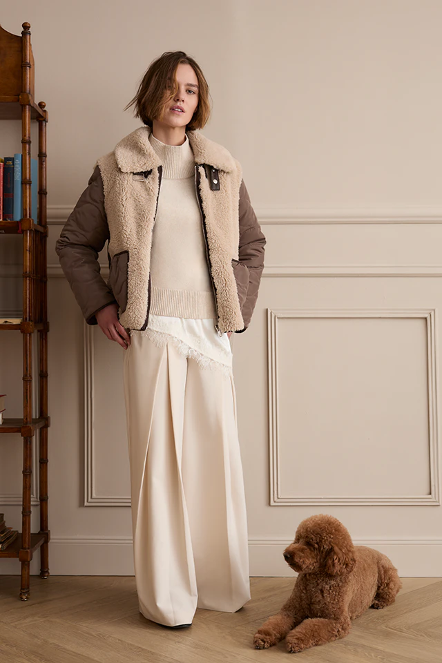 The model is wearing a brown jacket with a teddy bear lining over a cream-coloured knitted jumper and loose-fitting cream-coloured trousers. A brown dog is lying next to her.