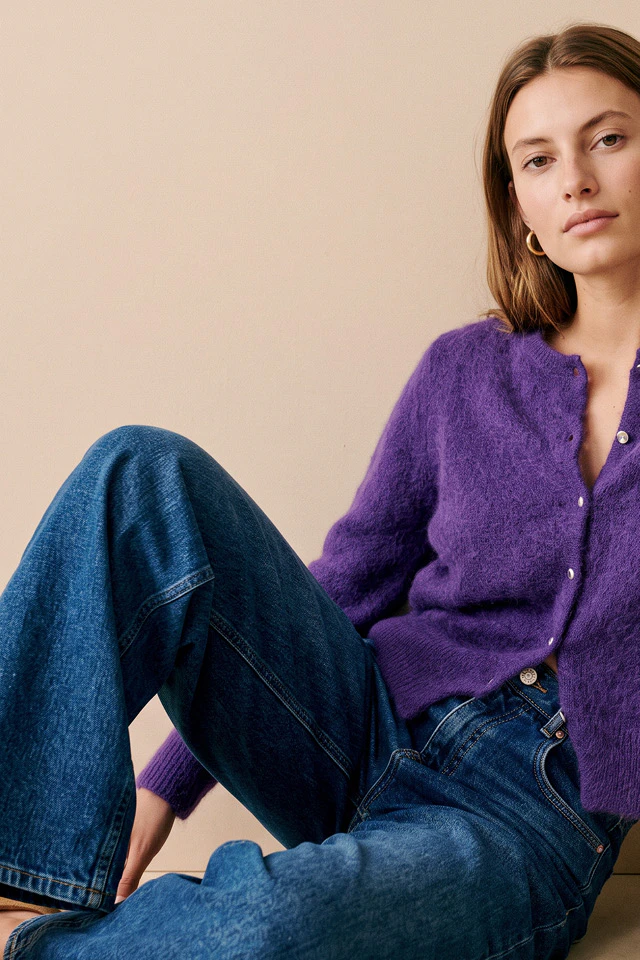 The model is wearing a fluffy purple cardigan with a button placket and dark blue jeans. She is sitting casually in front of a warm, beige background.
